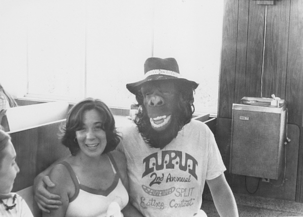 Black and white photograph. The room is a cafeteria seating area consistent with the 1970s or 1980s. There is a harsh glare in the background from the large window. A water cooler appears on a wall. A white person in a gorilla mask sits in the middle of the frame. There is a bucket hat on the person's head. The tee shirt worn says "IUPUI 2nd Annual Banana Split Eating Contest". The person in the mask has an arm around a laughing young white woman sitting very close. She has short dark hair and is wearing a