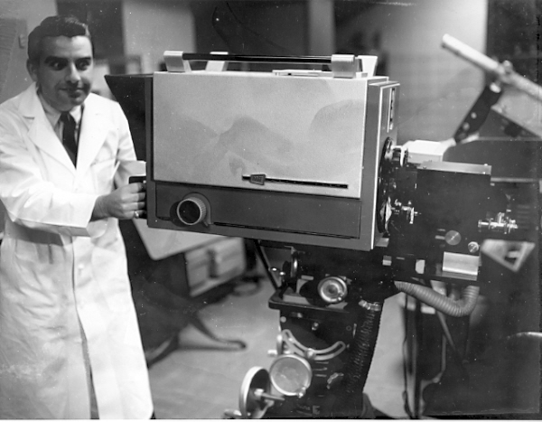 Black and white photograph. The year is 1975. The setting is a photography studio. Cameras and equipment consistent with that era decorate the room. A white man in a white lab coat is in the foreground on the left side of the frame. There is a ring on the pinky finger of his right hand. His face has an intense look, and his eyes are glaring at a large camera on a cart which he grips with his right hand.